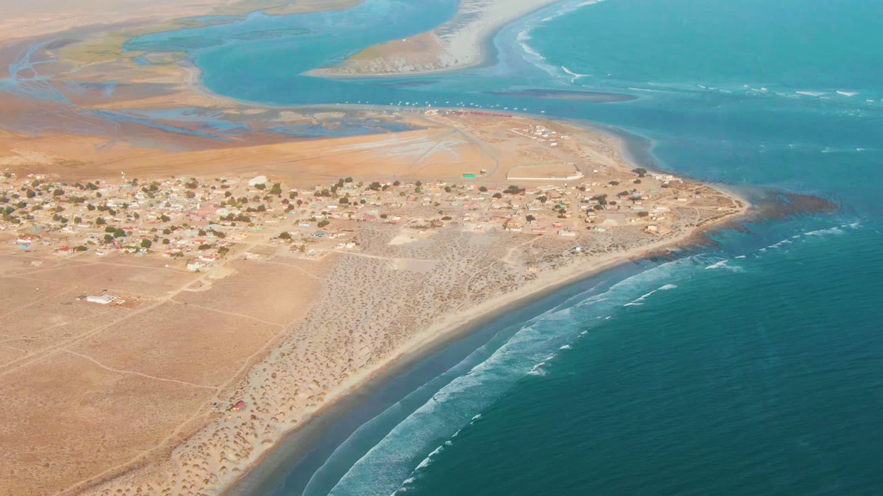 A high aerial frame over La Bocana, Baja California Sur shows a sandy village, curving beach, teal surf, distant sandbars, dry desert terrain, and no visible people.