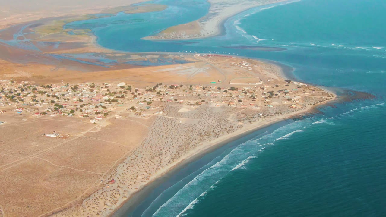 At 0:02, the drone frame shows La Bocana's settlement stretching along dry sand, a broad turquoise inlet beyond it, rolling white surf, and no visible people.