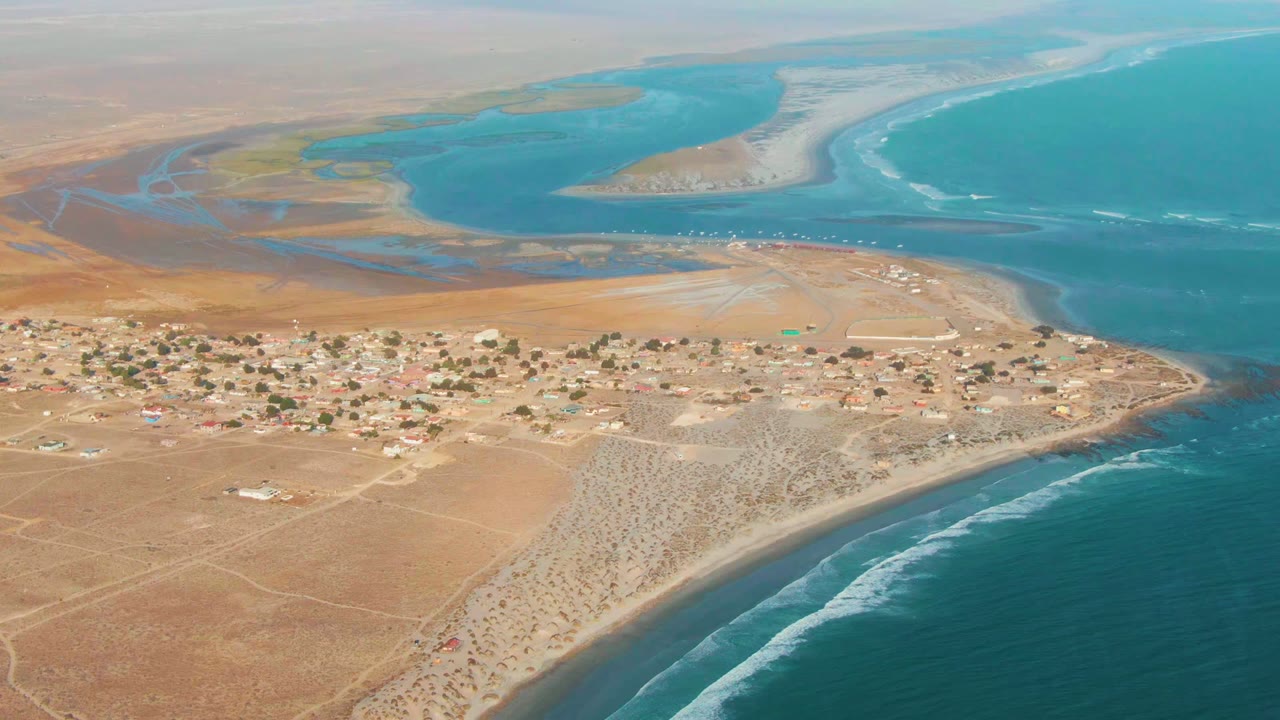 Wide aerial still over La Bocana showing clustered low buildings, pale sand, blue ocean, white wave lines, and lagoon water beyond the village, with no visible people.