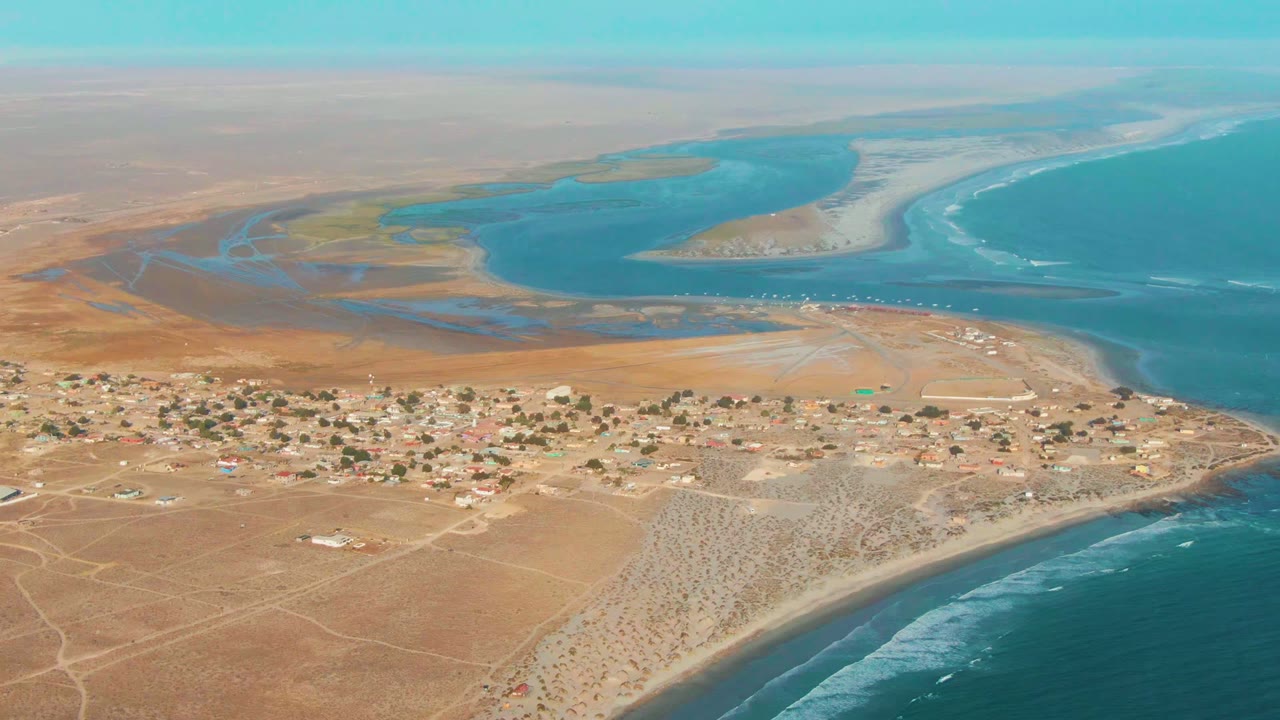Opening aerial frame of La Bocana, Baja California Sur, showing a small desert village between tan land, a lagoon, sandy beach, and ocean surf, with no visible people.