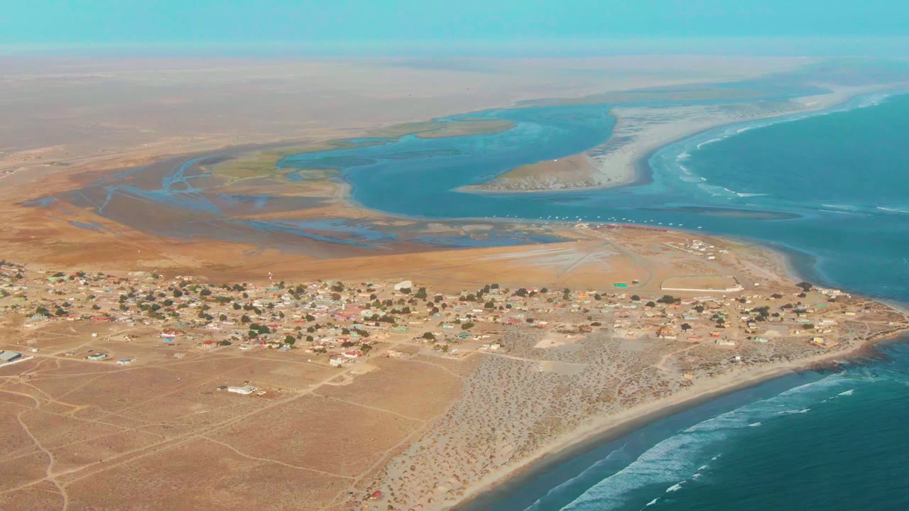 Aerial view over tan desert land, low village buildings, lagoon, and surf.