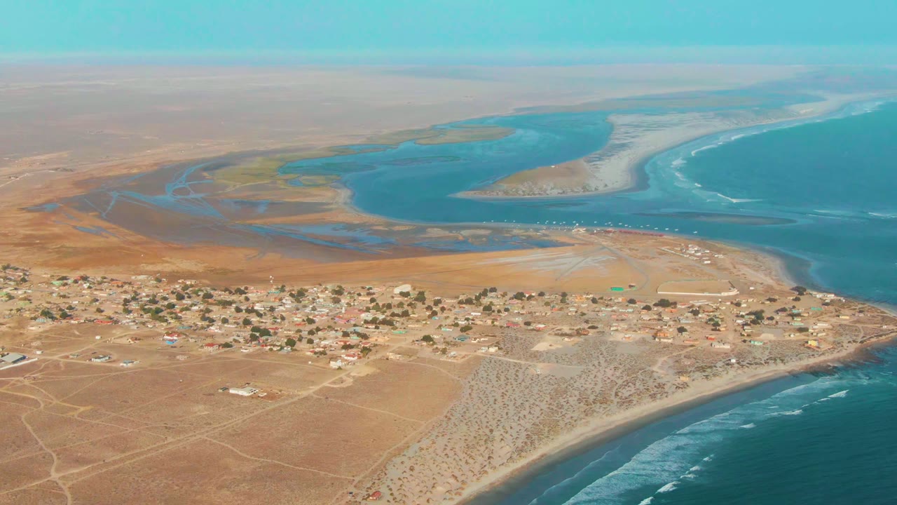 Third aerial frame of La Bocana frames the village along the lower center, lagoon water and sand flats in the background, and ocean surf wrapping the right shoreline, with no visible people.