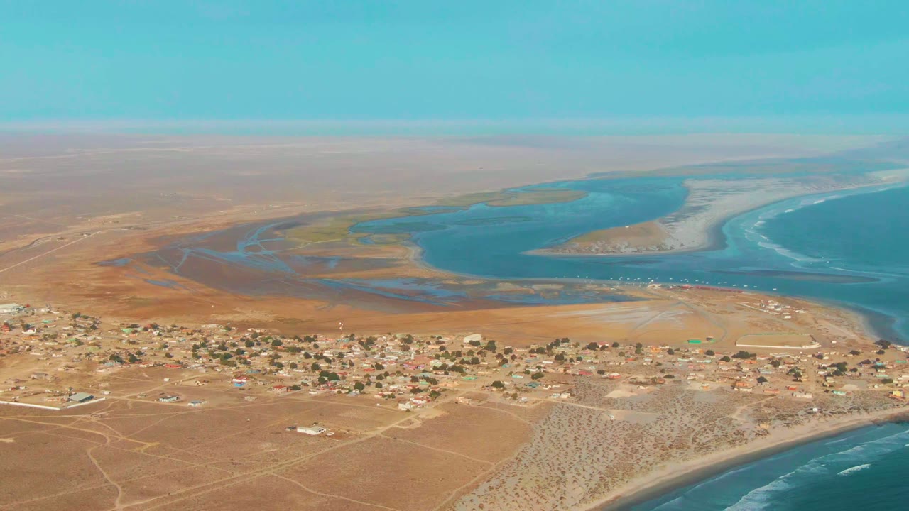 Opening aerial frame over La Bocana, Baja California Sur, with scattered buildings on brown sand, blue lagoon channels, surf on the right, and no visible people.