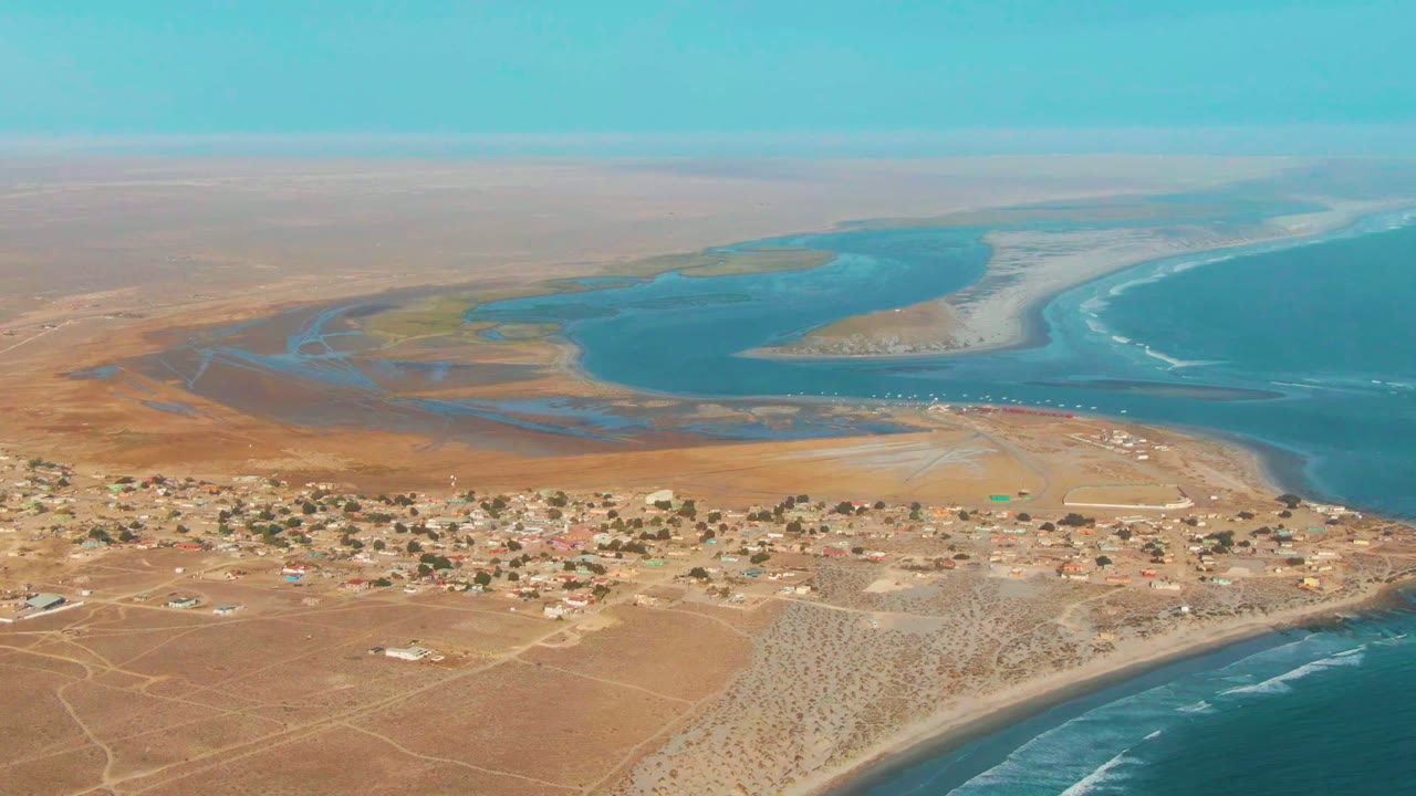 Opening aerial frame over La Bocana, Baja California Sur, with scattered low buildings in tan desert, blue lagoon water beyond them, ocean surf at right, and no visible people.