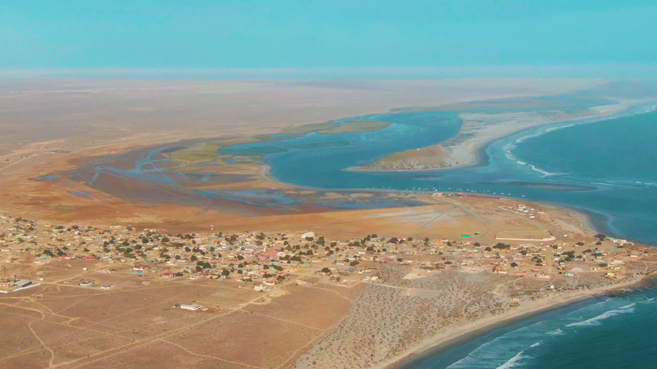 Wide aerial view of tan desert, a small town, blue lagoon, and ocean shoreline.
