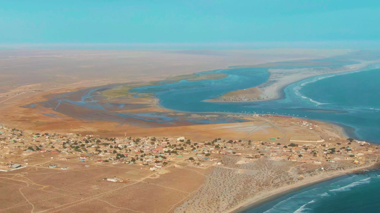 Aerial frame at 0:02 over La Bocana, Baja California Sur, with more open desert at left, clustered buildings below, blue lagoon water behind them, pale sandbars, and no visible people.