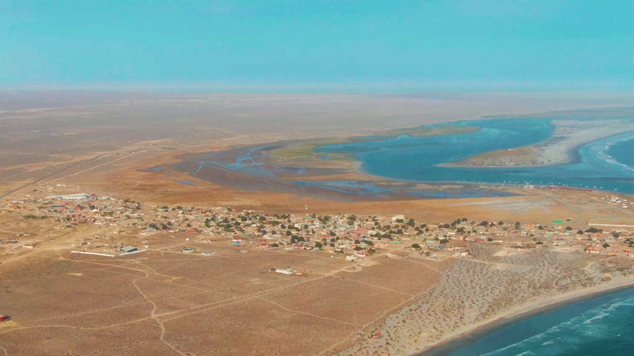 Opening aerial frame over La Bocana, Baja California Sur, showing tan desert ground, a scattered settlement, blue lagoon water, ocean edge, and no visible people.