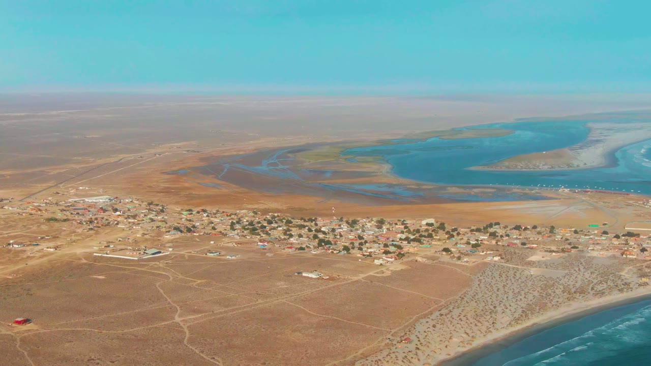 Aerial frame from La Bocana showing dry sandy ground, scattered homes, a turquoise lagoon, distant coast, surf at the lower right edge, and no visible people.