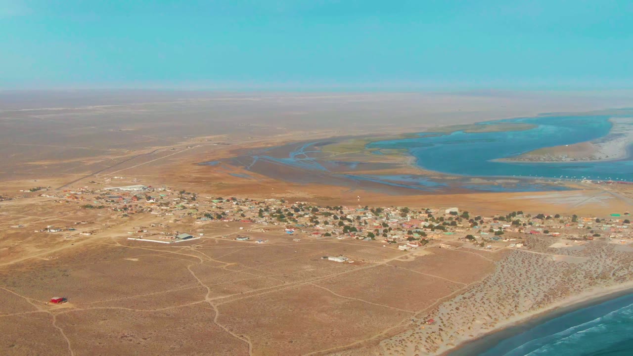 Aerial frame over La Bocana, Baja California Sur, showing a small desert village, dirt roads, ocean surf at the lower right, a blue lagoon beyond, and no visible people.