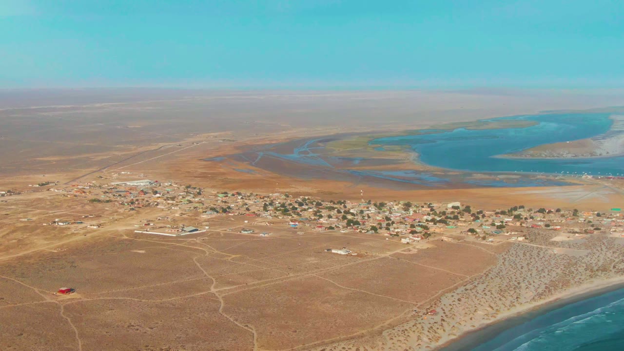High aerial frame of La Bocana showing the village stretched across brown arid ground, the lagoon occupying the upper right, ocean surf along the lower edge, and no visible people.