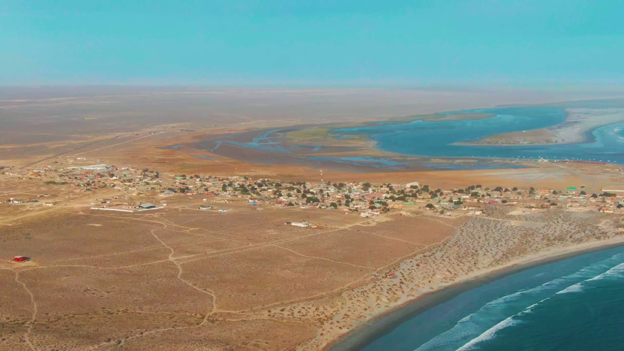 Wide aerial frame over La Bocana, Baja California Sur, showing a dry desert plain, scattered village buildings, beach surf in the foreground, blue lagoon water beyond, and no visible people.
