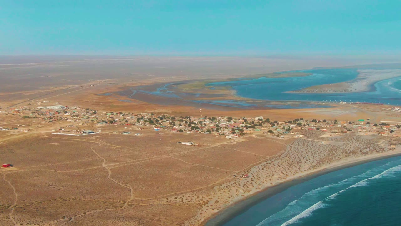 Aerial view of coastal desert roads, village buildings, beach, surf, and lagoon water.
