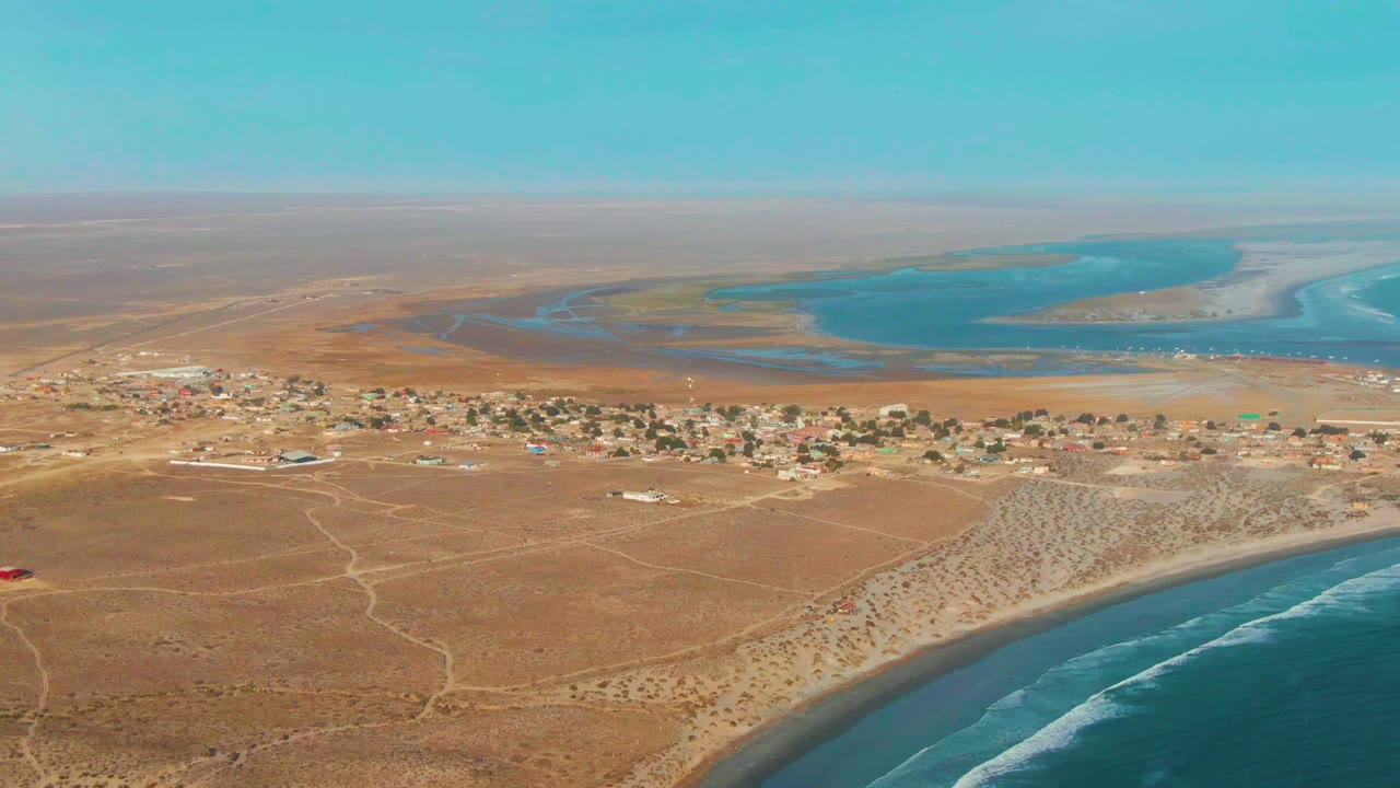 High aerial frame over La Bocana, Baja California Sur, with the settlement stretched across dry brown ground, ocean waves along the lower right beach, pale sandbars, and no visible people.