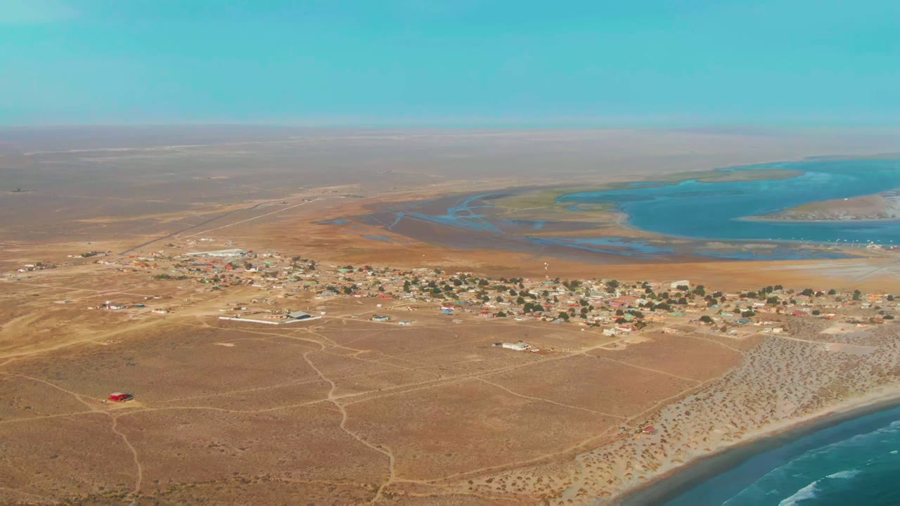 Opening aerial frame over La Bocana, Baja California Sur, with brown desert terrain, scattered village buildings, a blue lagoon in the distance, surf at the lower right, and no visible people.