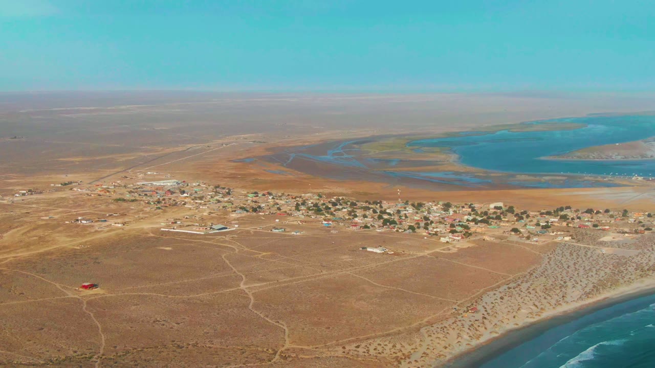 High aerial view of dry terrain, village buildings, lagoon water, and beach surf.