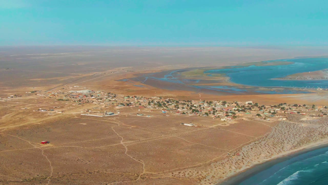 La Bocana appears from above with a broad brown foreground, faint dirt road lines, a compact village strip, lagoon water beyond the homes, ocean surf along the lower right edge, and no visible people.