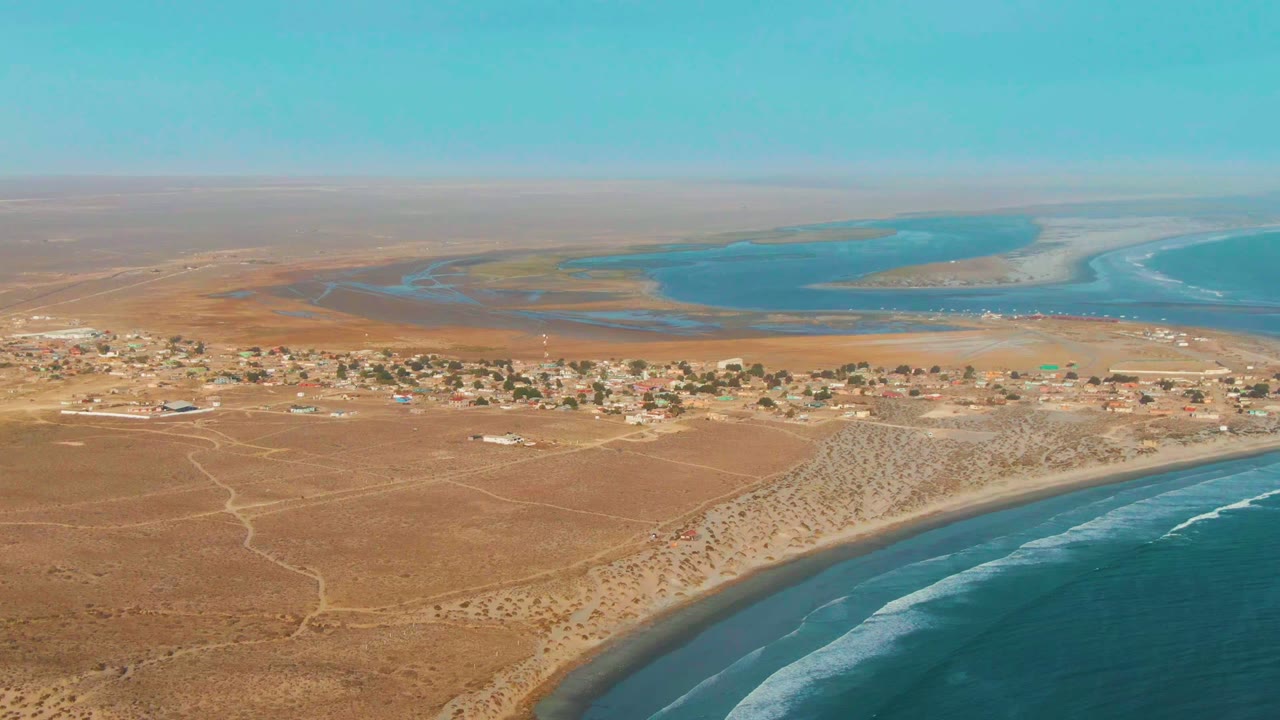 Wide aerial view over La Bocana, Baja California Sur, with sandy desert foreground, a small coastal village, curved beach, blue ocean, distant lagoon-like water, and no visible people.