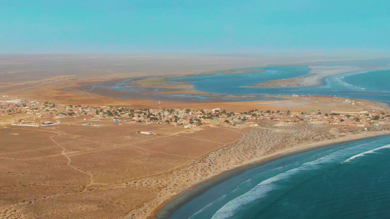 Drone view of sandy shoreline, scattered buildings, and turquoise coastal water