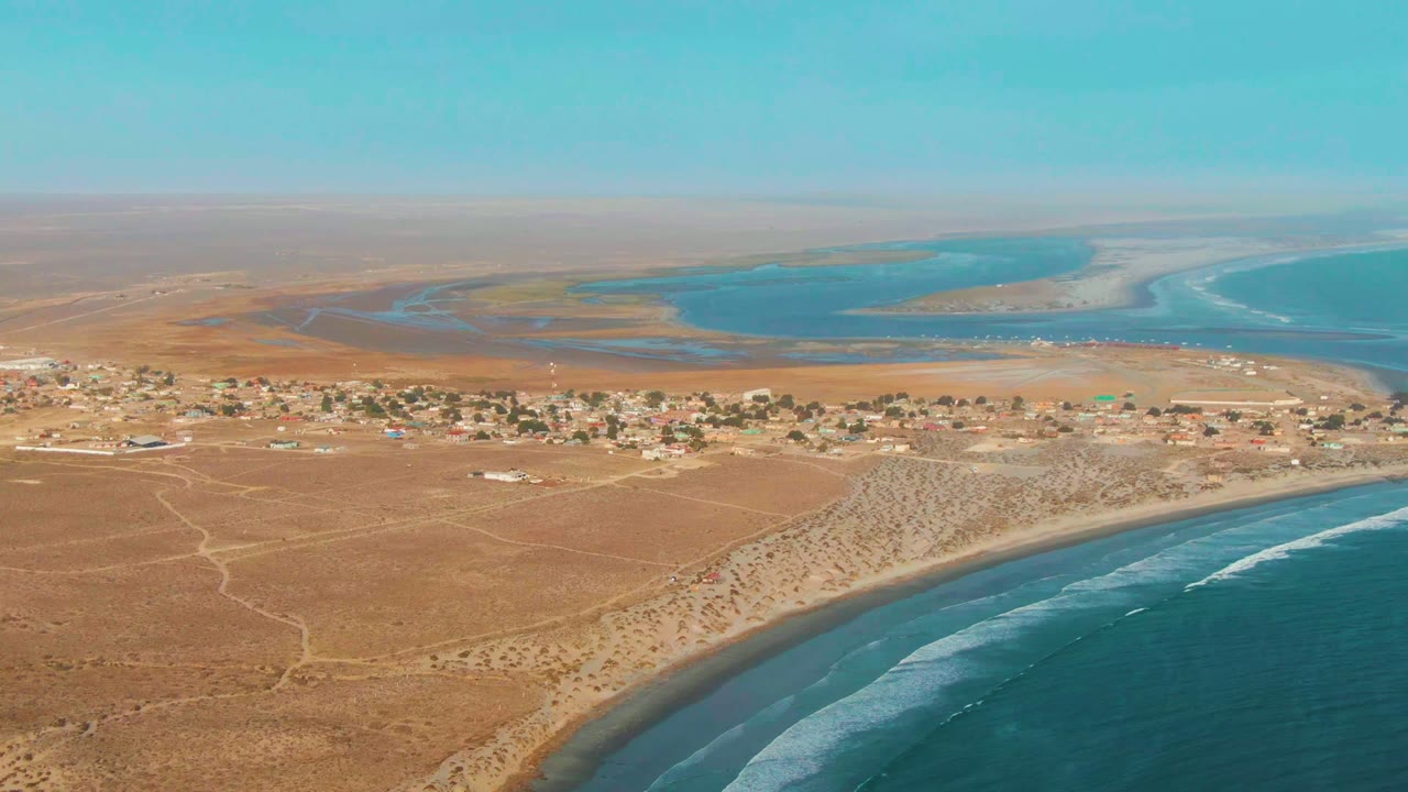 High aerial view of La Bocana with the village stretching across the midground, ocean surf along the lower right edge, inland blue water behind the settlement, and no visible people.