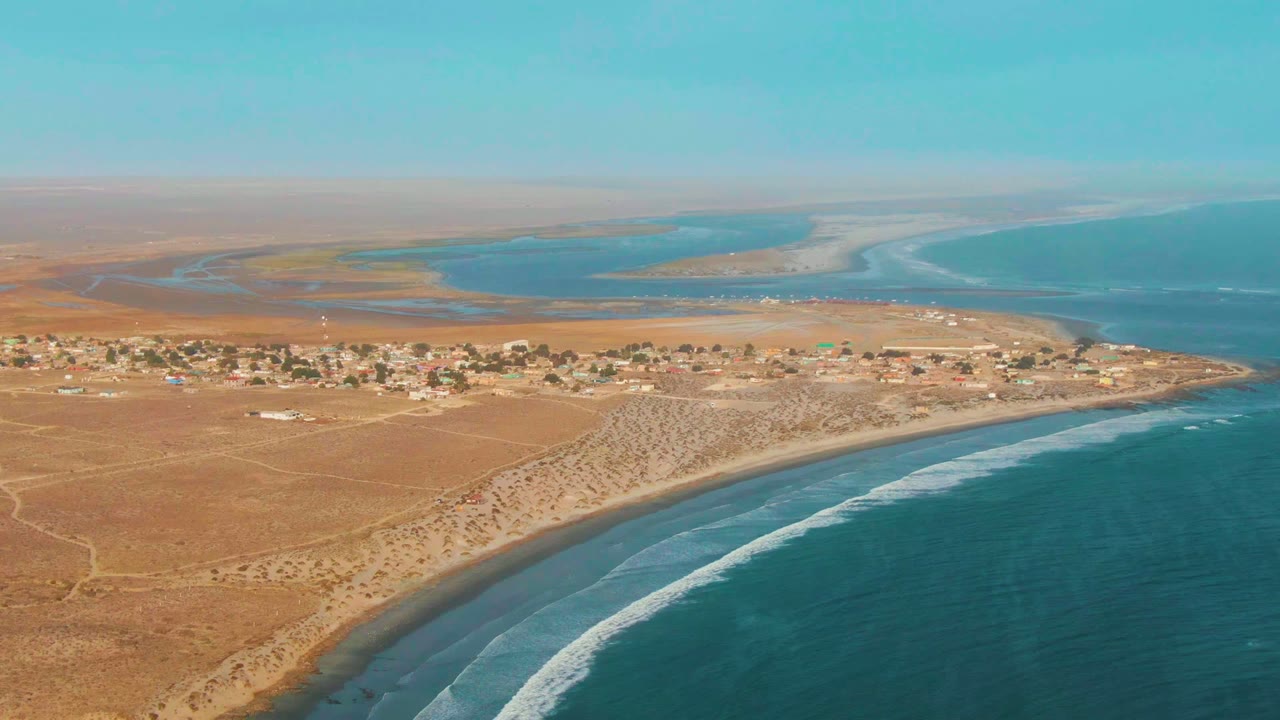 Aerial frame over La Bocana, Baja California Sur, showing a small village on arid ground above a curved beach with white surf and no visible people.