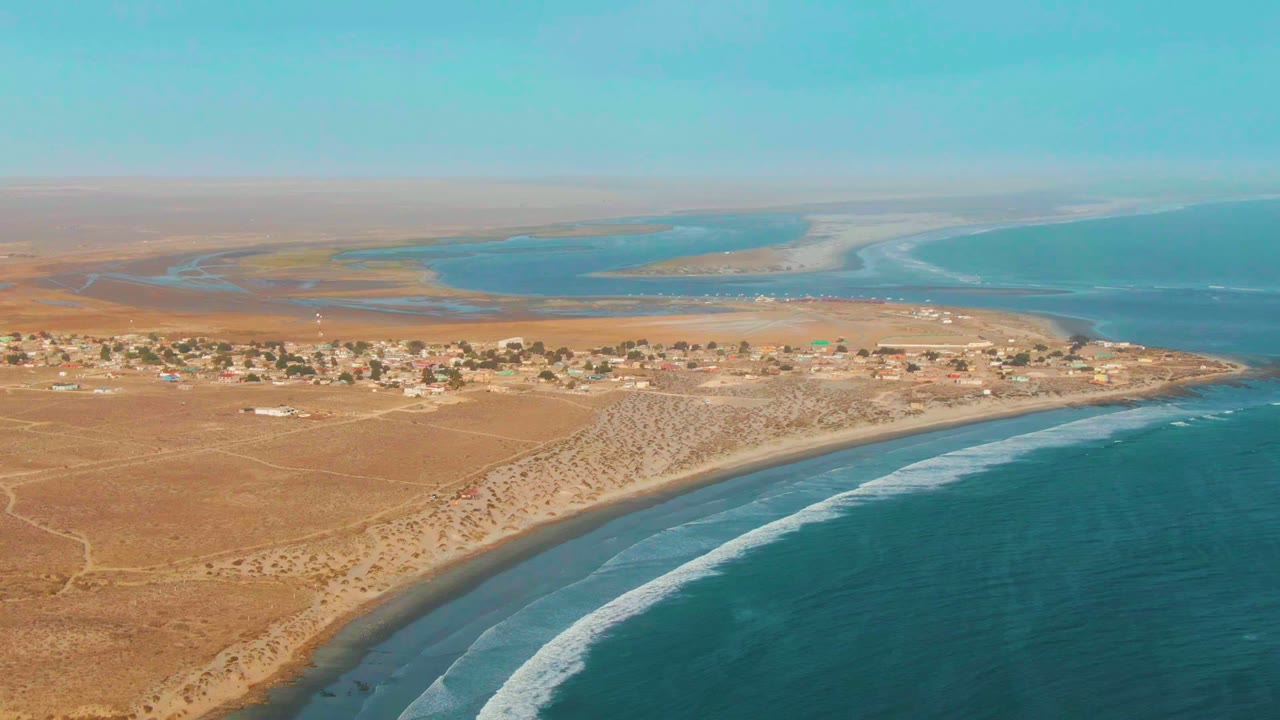 Wide aerial of dry coast, low buildings, beach, and ocean waves.