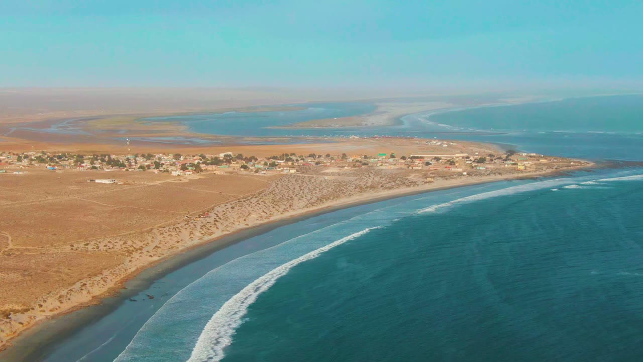 At 0:00, an aerial view over La Bocana shows tan desert land, a small coastal settlement, curved shoreline, blue ocean, white surf, and no visible people.