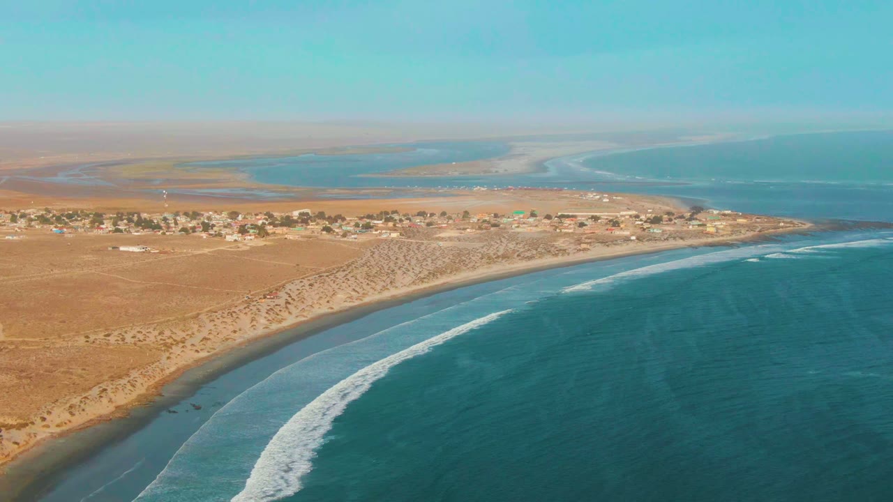 Wide drone view of sandy dunes, coastal buildings, and blue water.