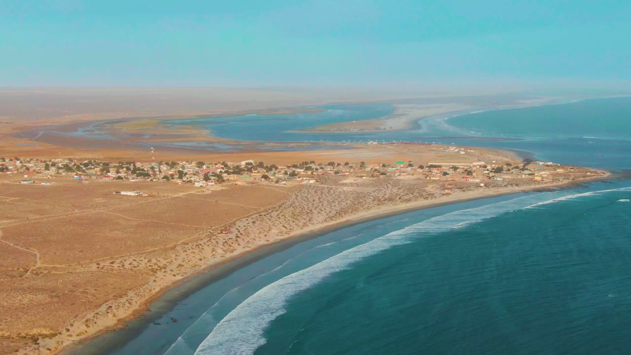 Opening aerial frame over La Bocana, Baja California Sur, with turquoise ocean in front, a curved sandy beach, tan desert settlement, distant lagoon channels, and no visible people.