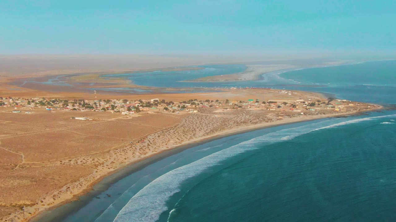 At 0:02, the drone frame holds the La Bocana coast with a broad band of blue ocean, white waves along the sandy edge, scattered settlement structures, dry terrain, lagoon water, and no visible people.