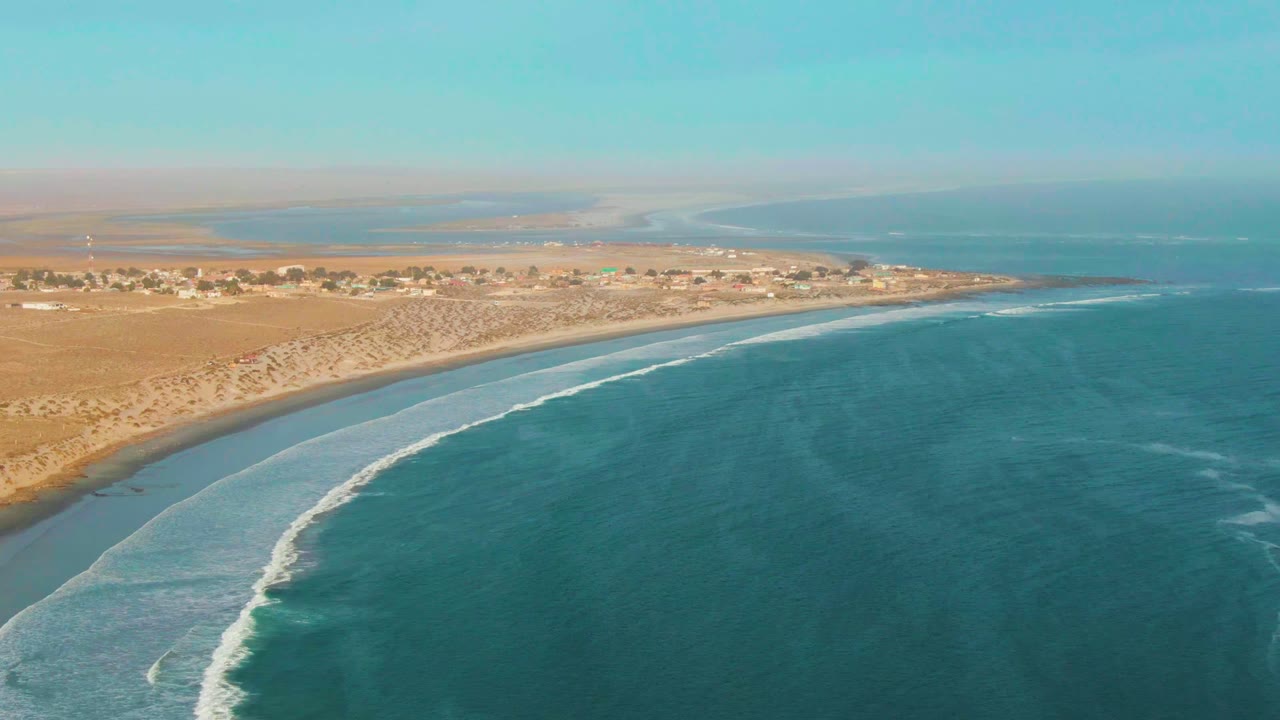 Aerial view over La Bocana, Baja California Sur, with blue-green ocean in the foreground, white surf curving toward a sandy beach, desert land and a small shoreline settlement in the distance, and no visible people.