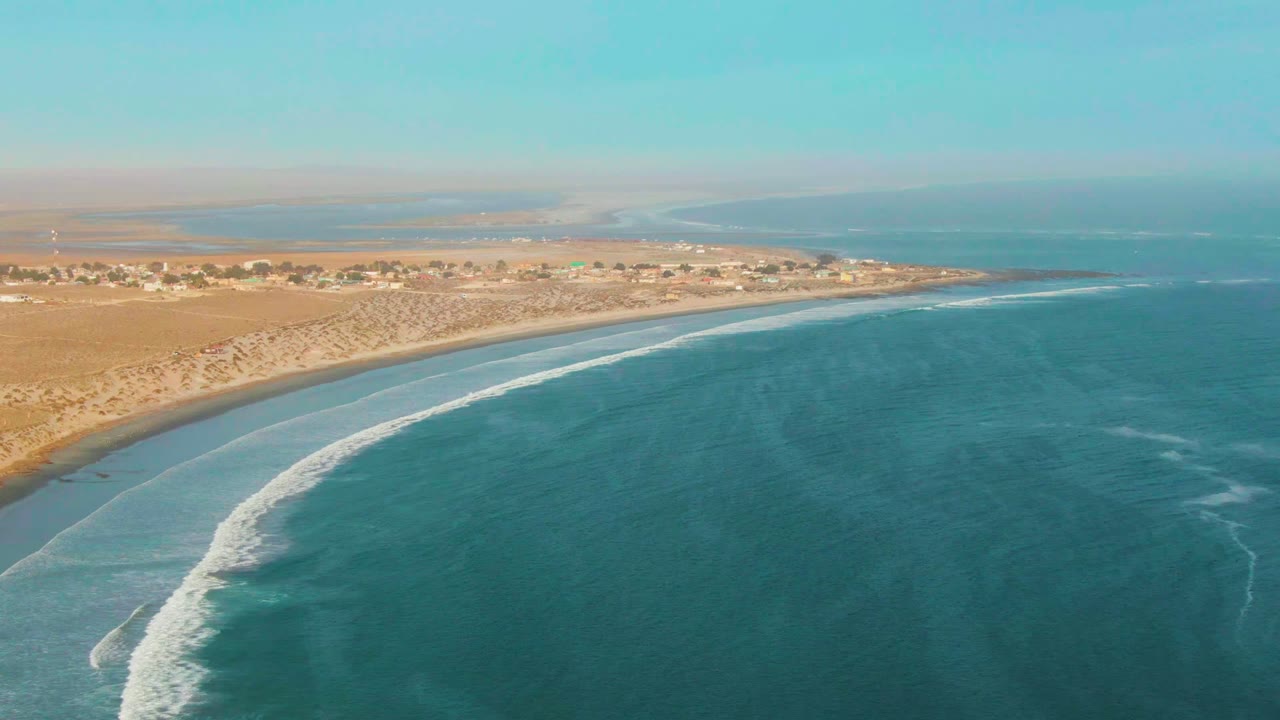 Wide aerial view of La Bocana with the surf line stretched diagonally through the frame, turquoise water to the right, sandy coast and desert settlement to the left, and no visible people.