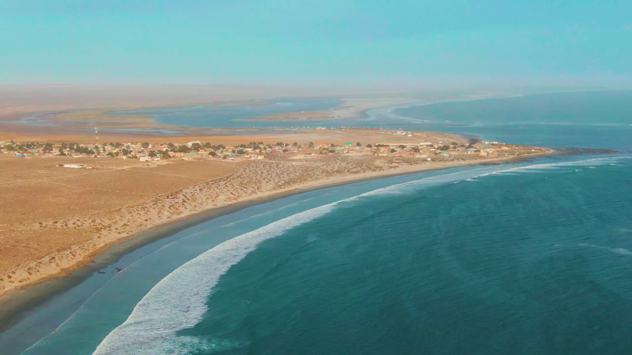Aerial view over La Bocana, Baja California Sur, with blue ocean on the right, a curved sandy beach below, low dunes and scattered buildings beyond, and no visible people.