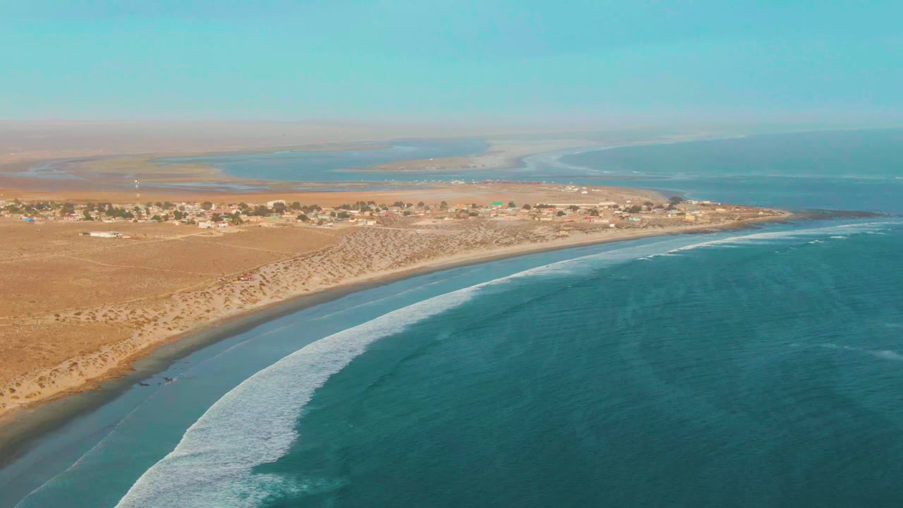 High aerial coastal view with white surf bending along sandy shore and desert settlement.