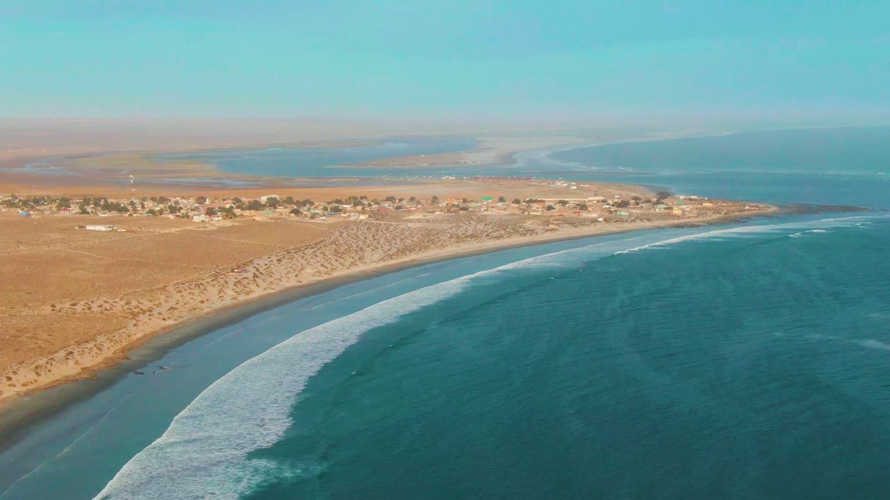 Drone-like aerial view of La Bocana with the wave line shifted farther along the curved shore, a dark wet-sand edge near the beach, inland dunes, small structures, and no visible people.