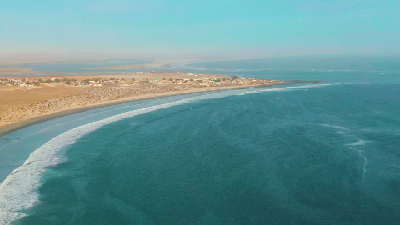 Aerial view over La Bocana, Baja California Sur, with turquoise water in the foreground, a curved band of white surf at the sandy shoreline, distant coastal buildings, and no visible people.