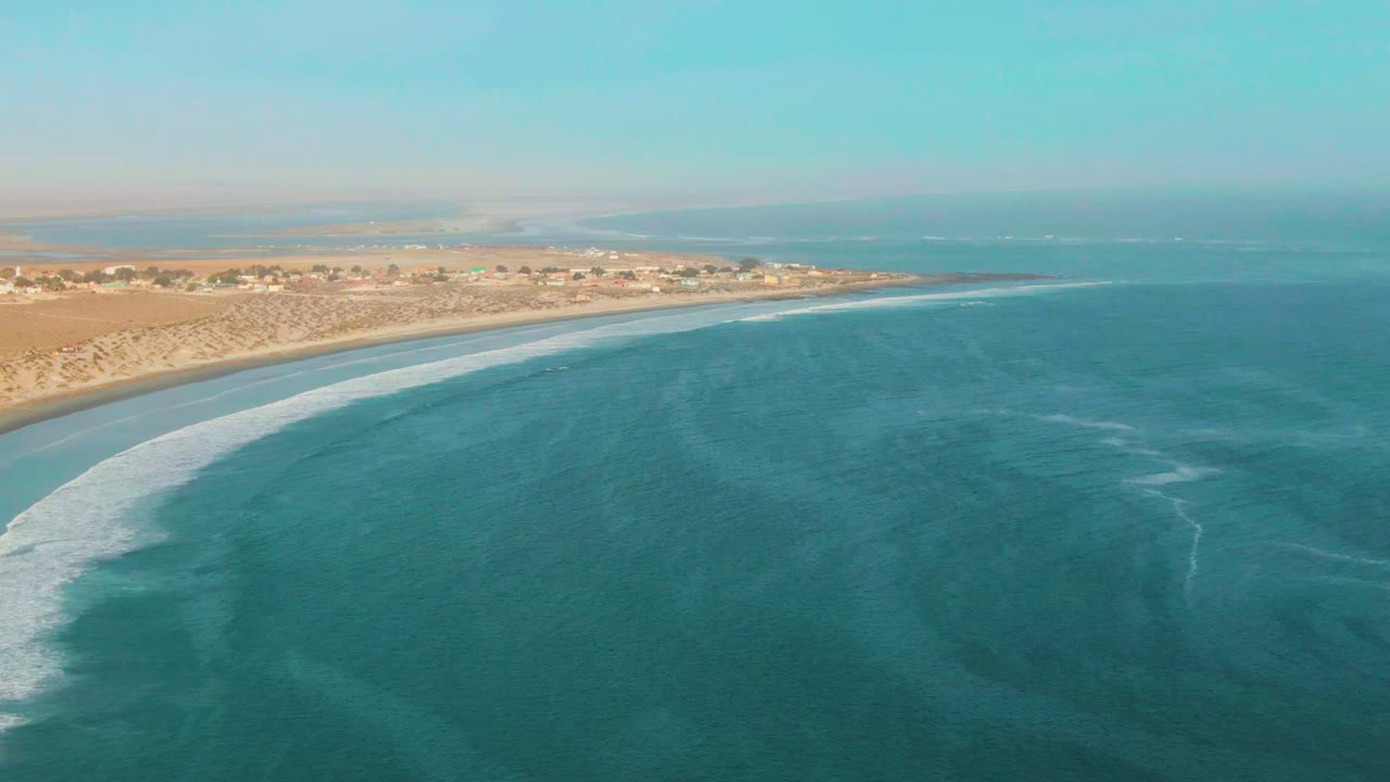 High aerial frame of La Bocana with open turquoise sea dominating the foreground, a white wave line touching the sand, a thin strip of coastal buildings, and no visible people.
