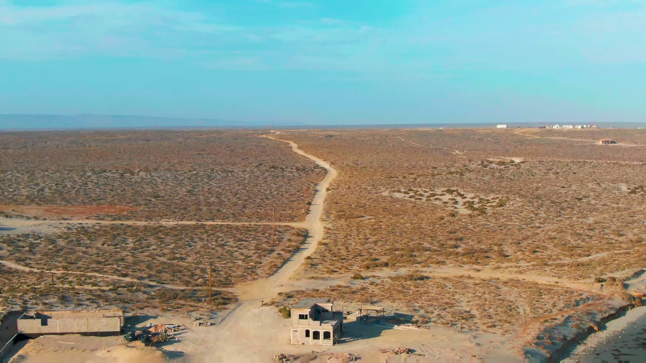 Aerial frame over La Bocana desert terrain showing a winding dirt road, two small abandoned structures, scrub vegetation, distant hills, clear blue sky, and no visible people.