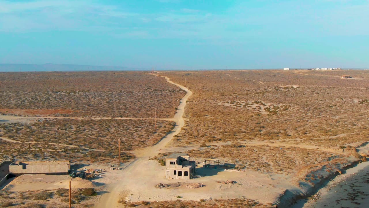 La Bocana desert frame with a broad sandy road crossing the foreground, low abandoned structures at lower right, sparse brush across the plain, and no visible people.