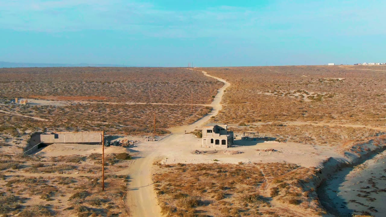Opening aerial frame over La Bocana, Baja California Sur, showing a pale dirt road, abandoned concrete structures, scrub desert, utility poles, blue sky, and no visible people.