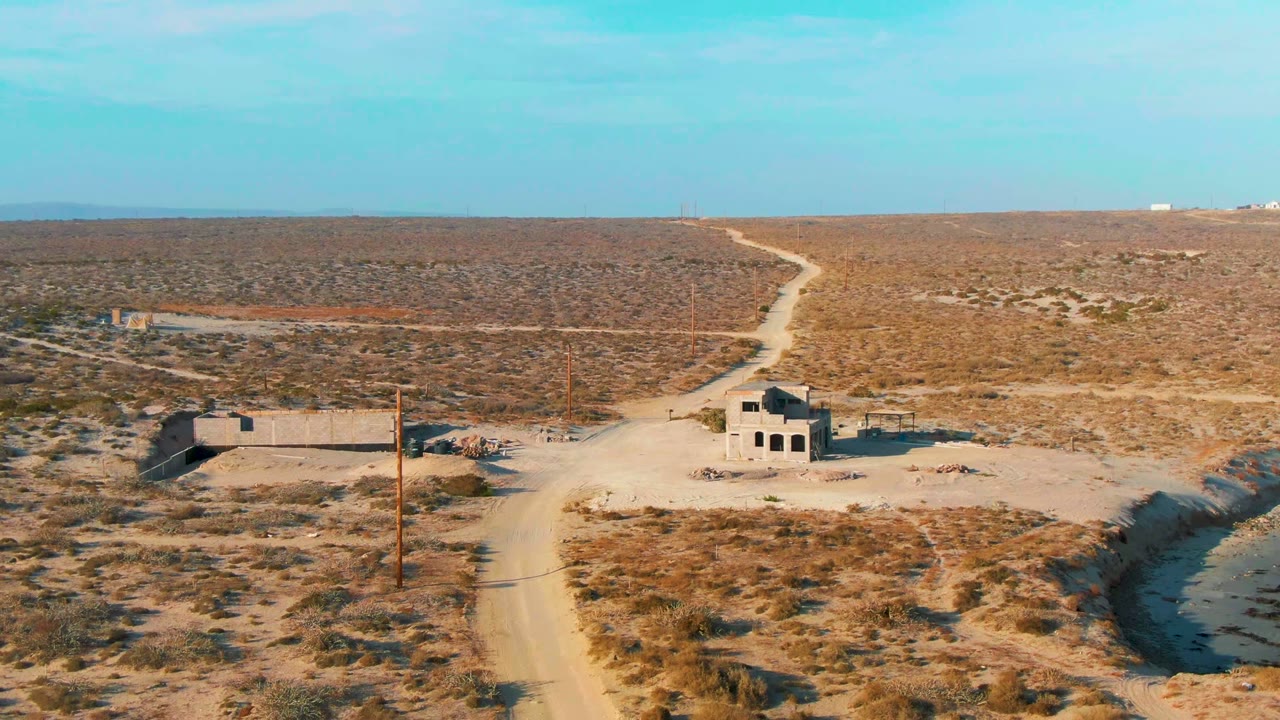 Mid-clip aerial frame over La Bocana, Baja California Sur, showing unfinished or abandoned buildings near a dusty road, open scrubland, a shallow sandy channel, and no visible people.
