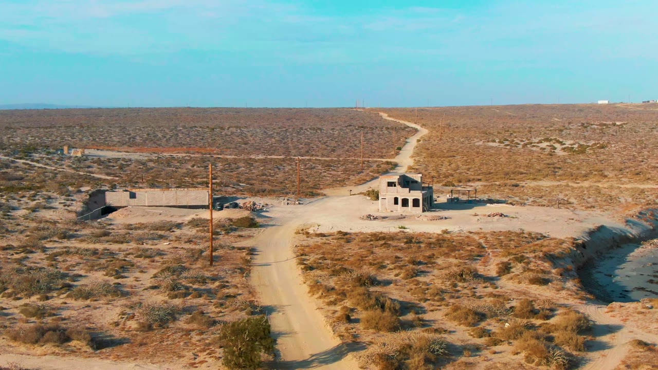 Aerial view in La Bocana, Baja California Sur, showing a sandy dirt road leading past abandoned buildings, utility poles, sparse shrubs, and a flat desert horizon with no visible people.