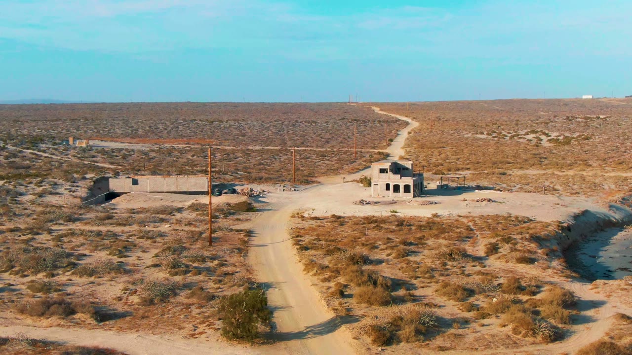 Dirt road curves through dry scrub toward small abandoned structures under a blue sky.