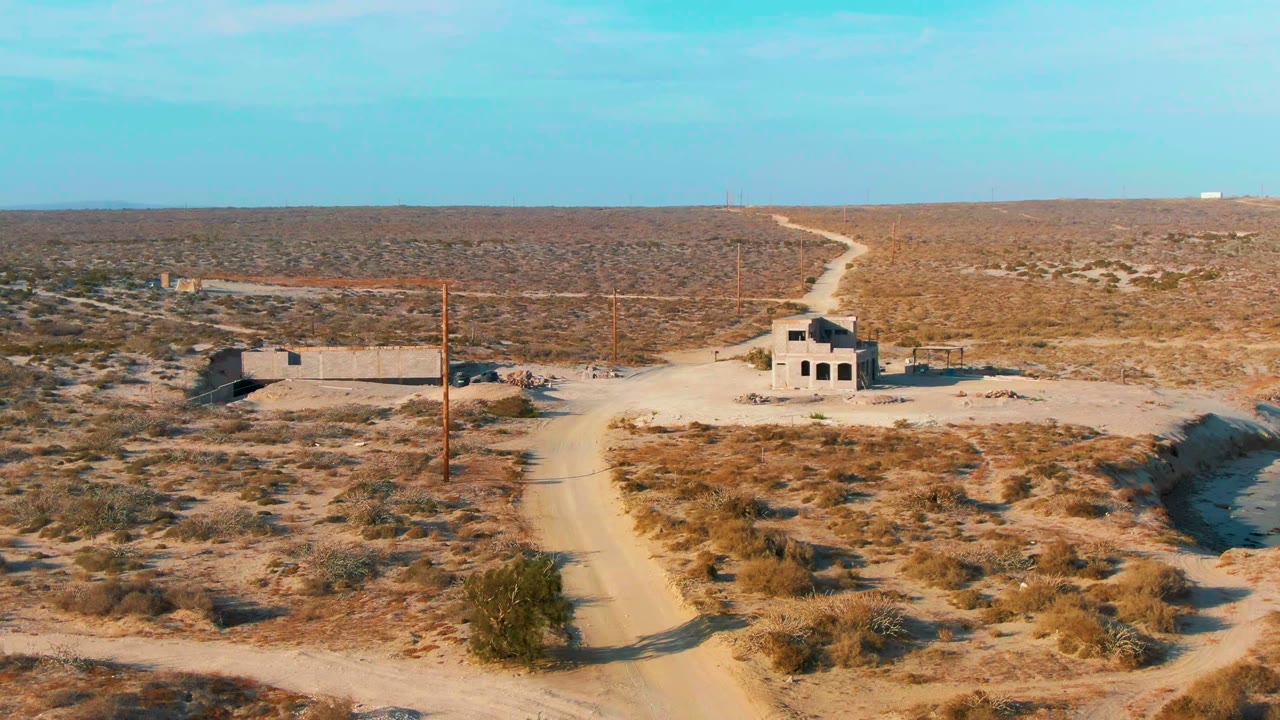 A wide aerial frame over La Bocana shows the dirt road crossing the foreground toward abandoned buildings and utility poles, backed by open desert and no visible people.