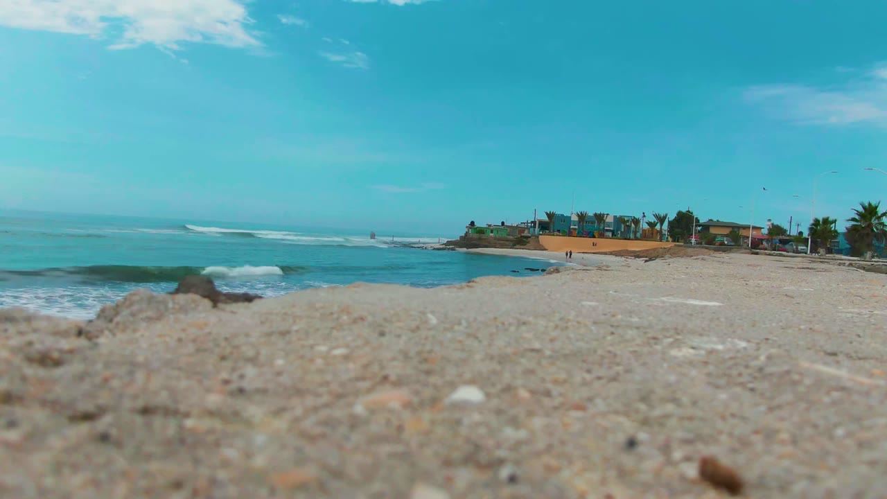 Low sand-level view at Punta Abreojos, Baja California Sur, with turquoise ocean, a breaking wave, distant shoreline buildings, palm trees, blue sky, and no visible people.