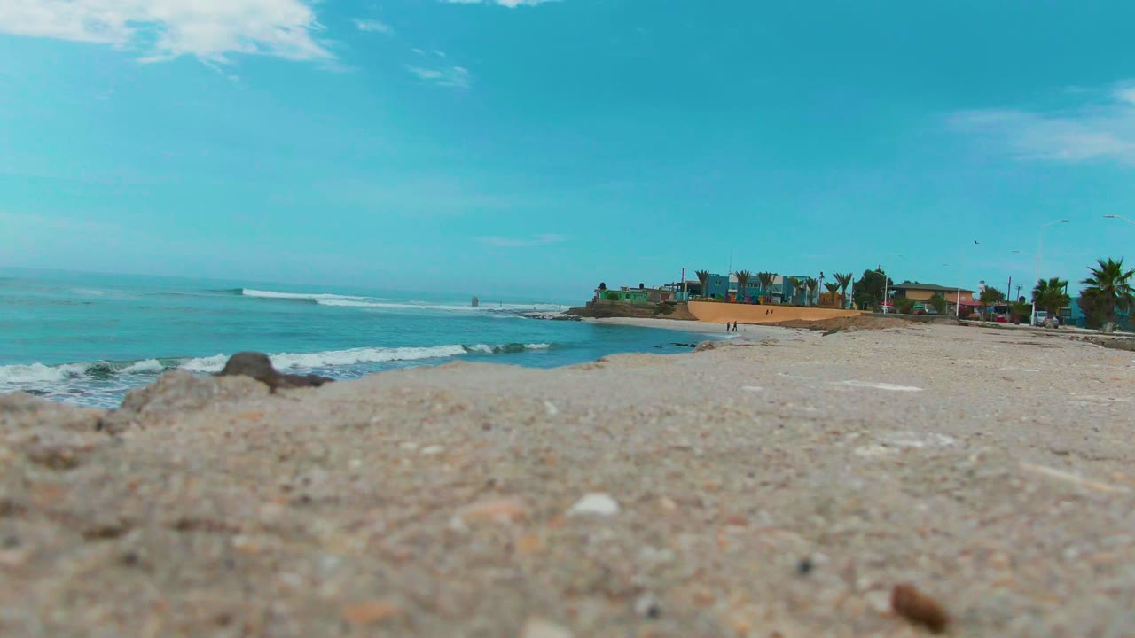 A low Punta Abreojos shoreline frame shows blurred sand in front, calm turquoise water, white wave foam, coastal buildings, palms, and blue sky with no visible people.