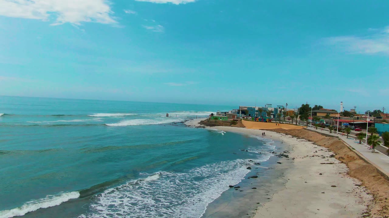 Aerial view over Punta Abreojos, Mexico, with blue ocean waves curling toward a sandy and rocky beach, small coastal buildings in the distance, and tiny people visible near the shoreline.