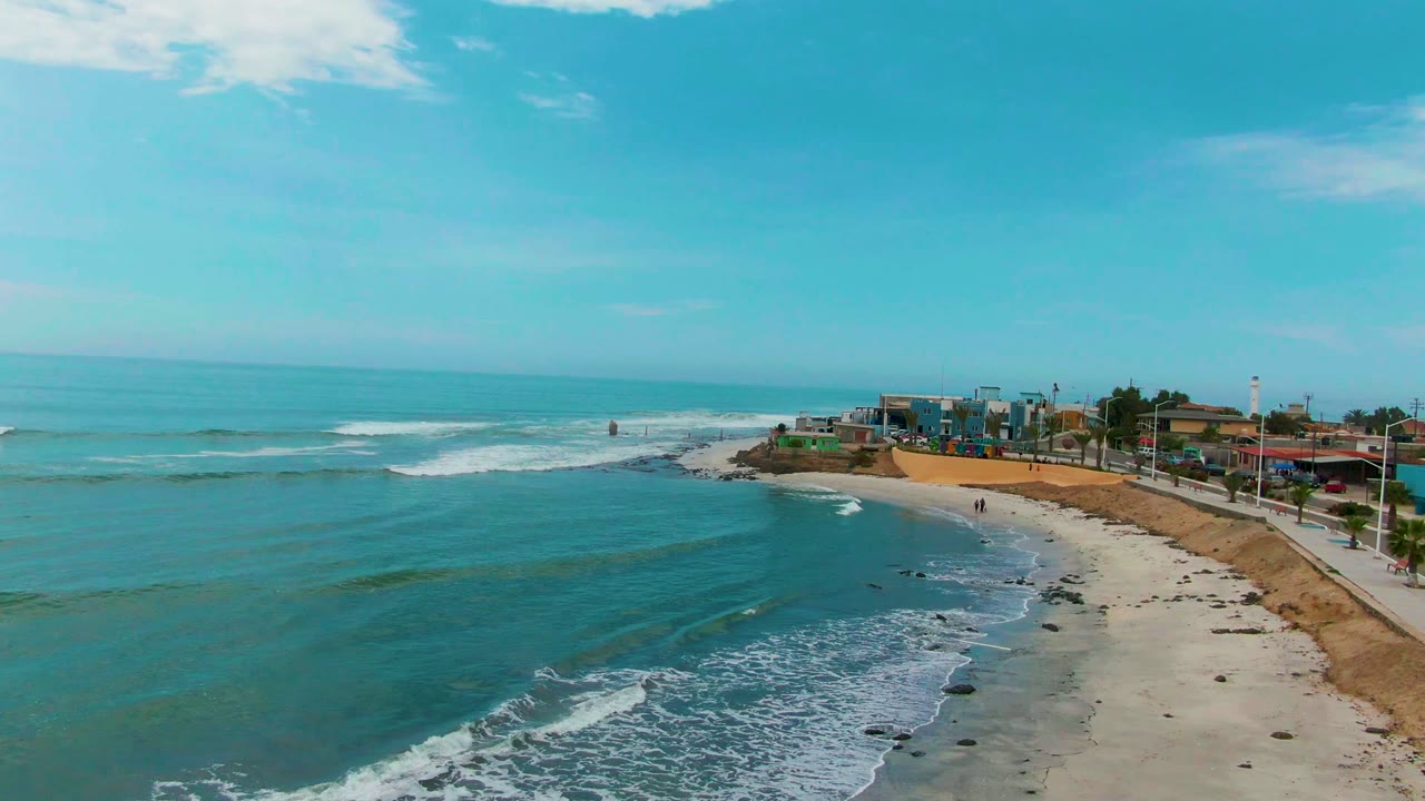 Wide coastal view with low waves, sandy shore, and buildings beyond the beach.