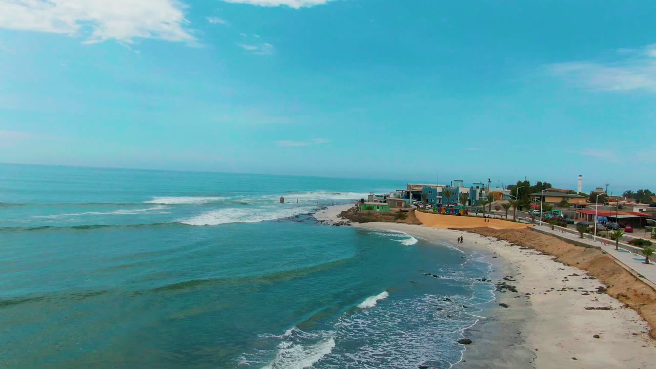 Aerial view of Punta Abreojos with a larger expanse of blue-green water in front, a narrow sandy beach to the right, low buildings along the shore, and small walkers visible near the water.