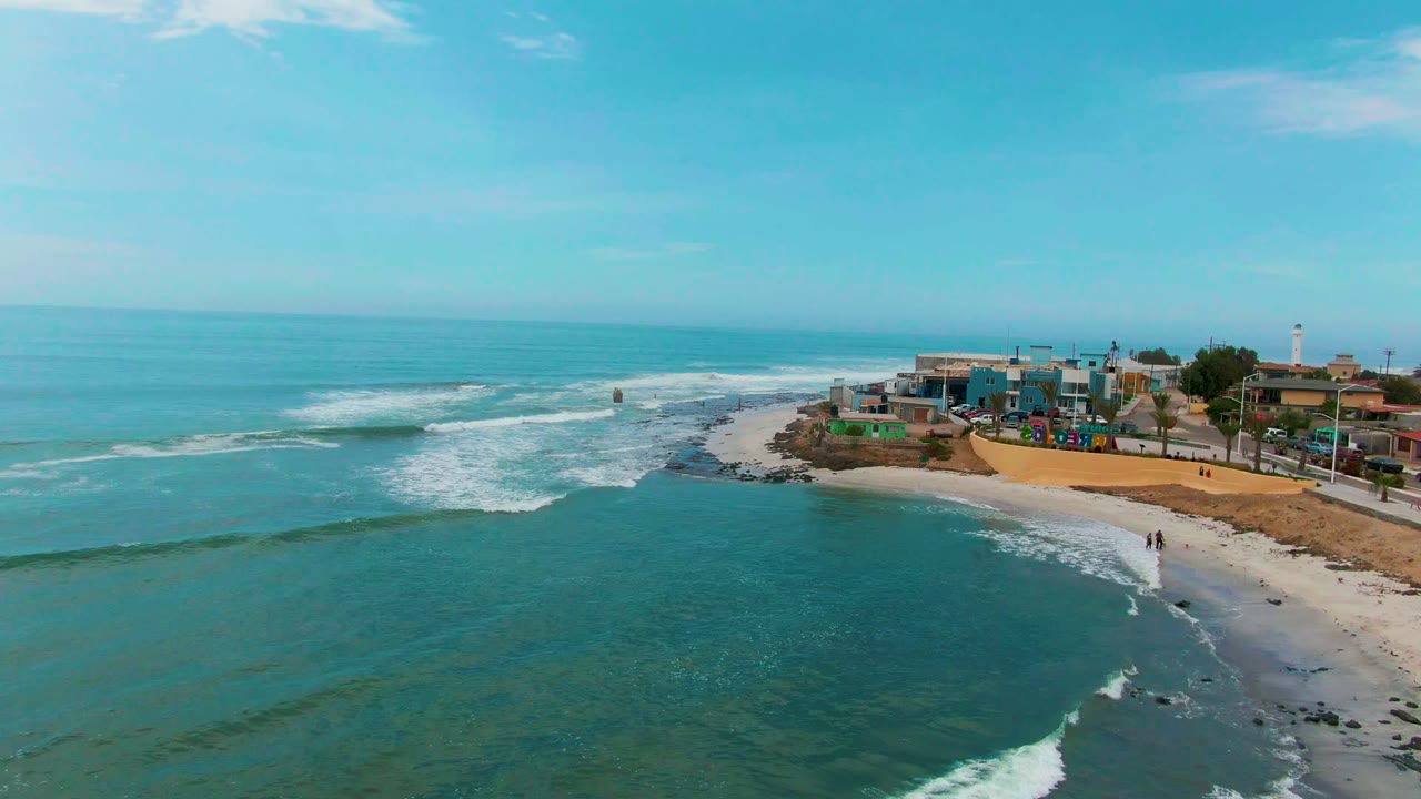 Aerial view in Punta Abreojos, Baja California Sur, with blue-green water in the foreground, white surf along the beach, coastal buildings to the right, and a few distant people on the sand.