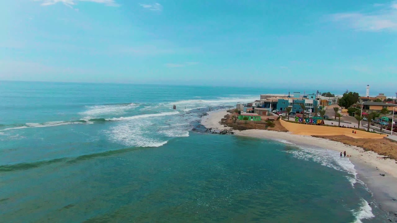 Foamy waves approach a sandy shoreline with buildings along the right edge.